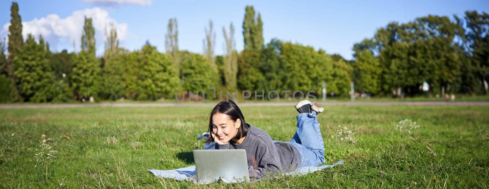 Portrait of young happy asian girl lying on blanket in park, watching videos and browsing internet on her laptop.