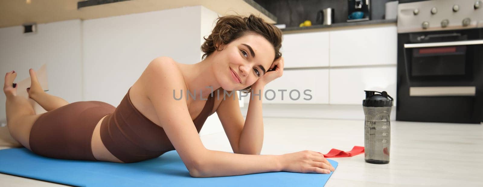 Sport and healthy lifestyle. Portrait of brunette sportswoman doing fitness in activewear, lying on rubber mat on floor, doing workout training in kitchen, drinking water from gym bottle by Benzoix
