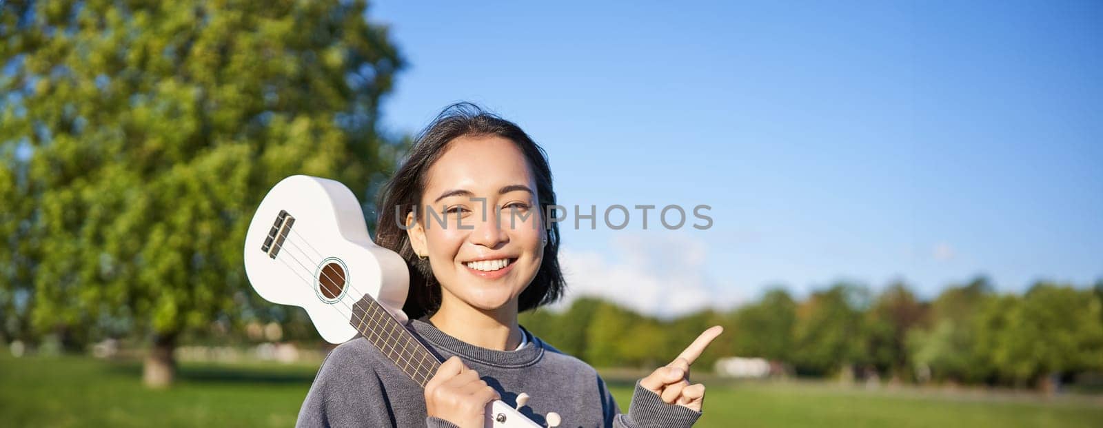 Portrait of asian smiling girl, holding ukulele over shoulder, pointing finger at copy space, banner or logo.