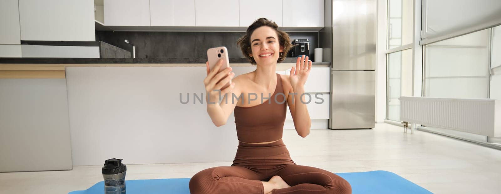 Smiling fitness girl, young athlete sits at home on yoga mat, waves hand at smartphone, joing online workout training session from home, says hello by Benzoix