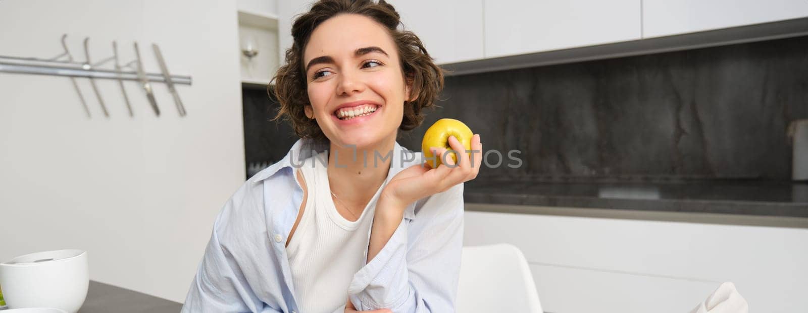 Image of brunette woman laughing, smiling while eating an apple in kitchen, sitting at home, having a snack. People and lifestyle concept