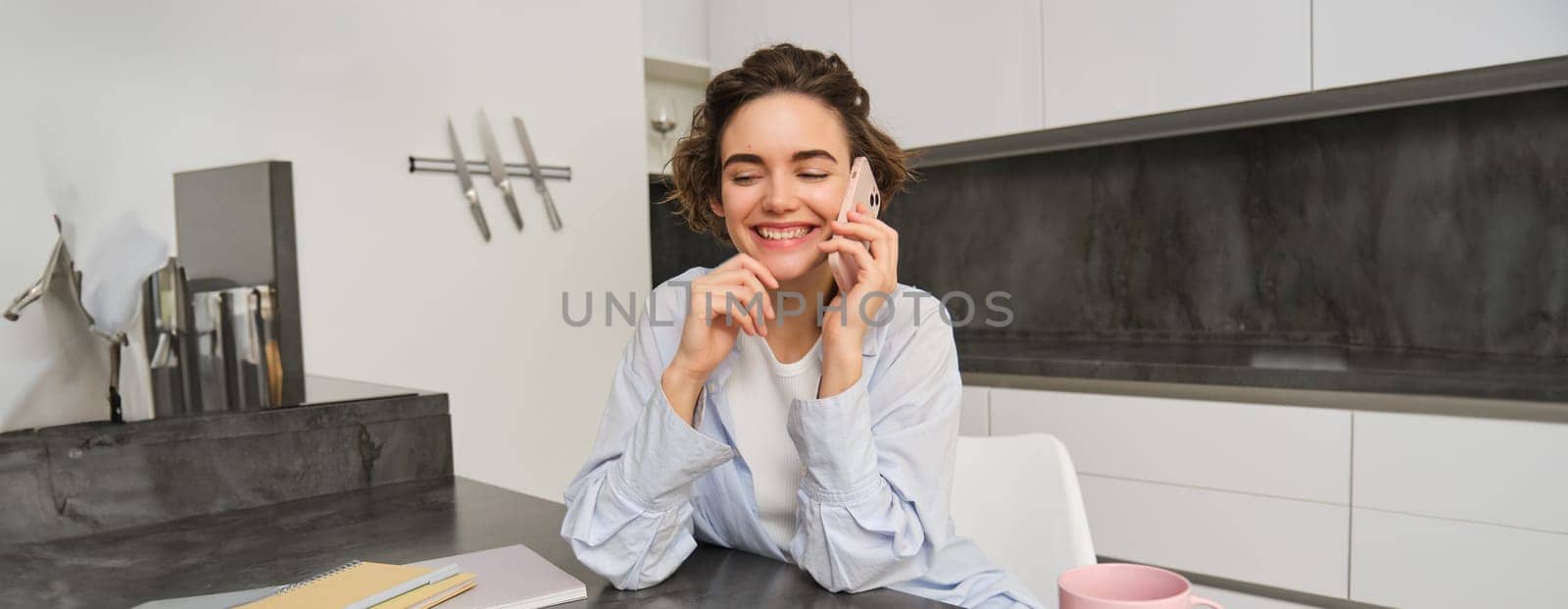Smiling young woman talks on mobile phone, calls someone from home, sits in kitchen and has conversation. Copy space