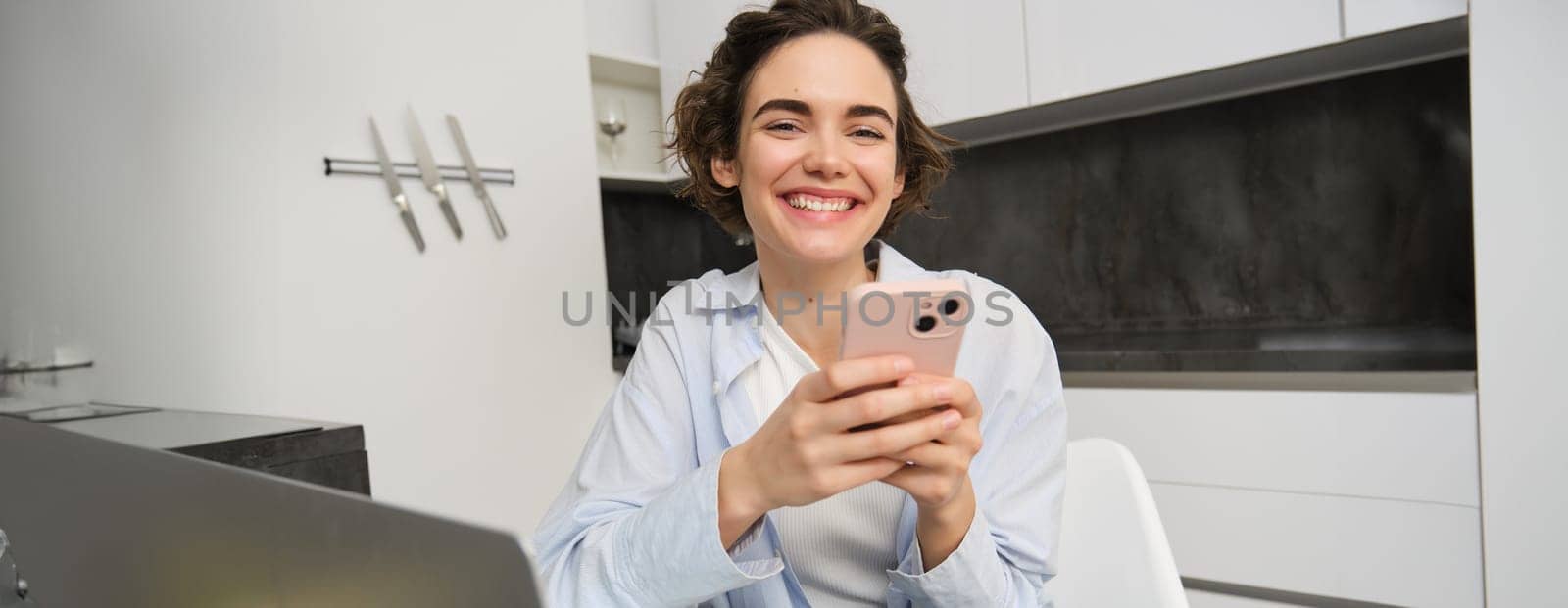 Image of young modern woman, holding mobile phone, sitting in her kitchen at home, order delivery on smartphone app.