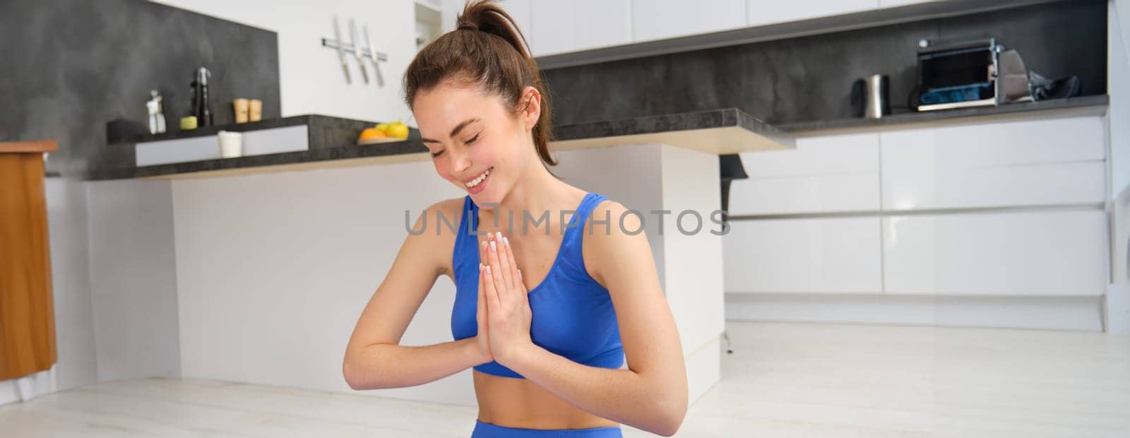 Portrait of smiling young yoga instructor, sitting at home in lotus pose, shows namaste gesture, meditating, doing online fitness class indoors by Benzoix