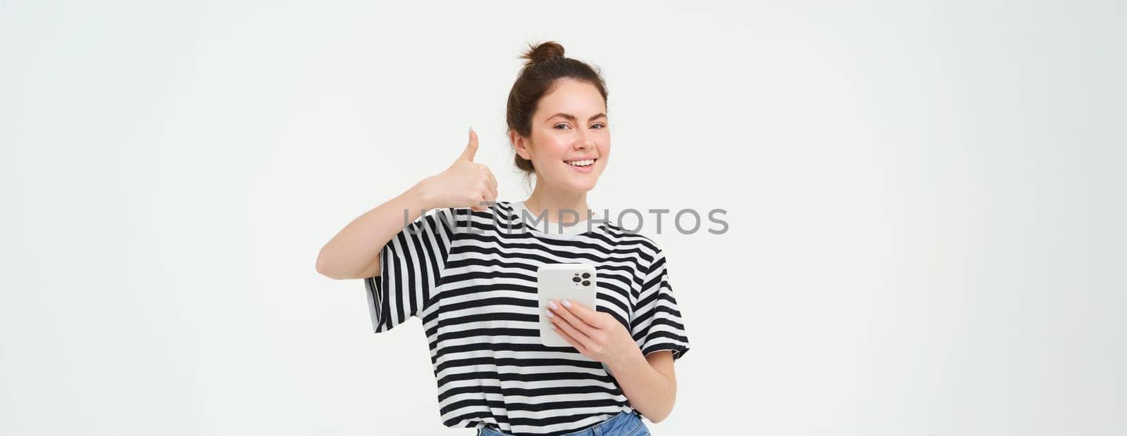 Cheerful, smiling young woman shows thumbs up, approves and recommends mobile app, holds smartphone, looks satisfied, stands over white background.