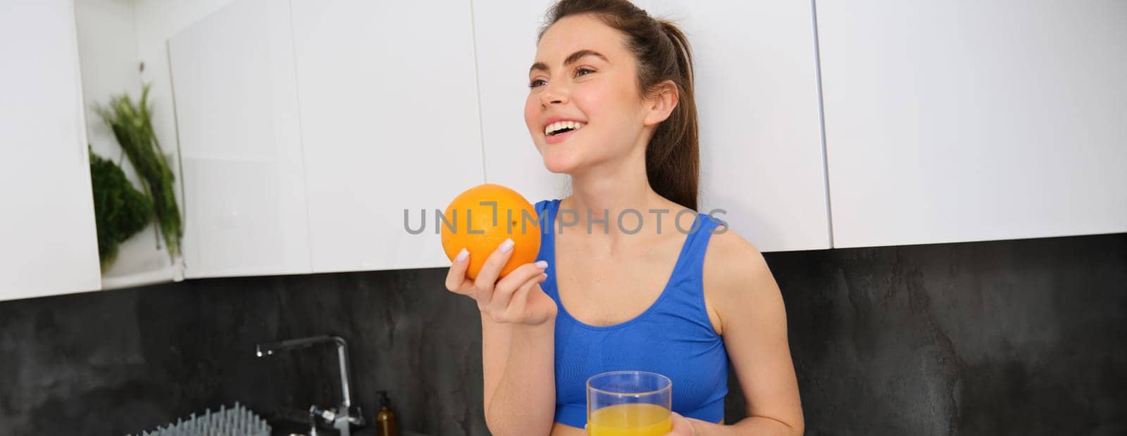Indoor shot of woman after workout, standing in kitchen with fresh juice and an orange, drinking it by Benzoix