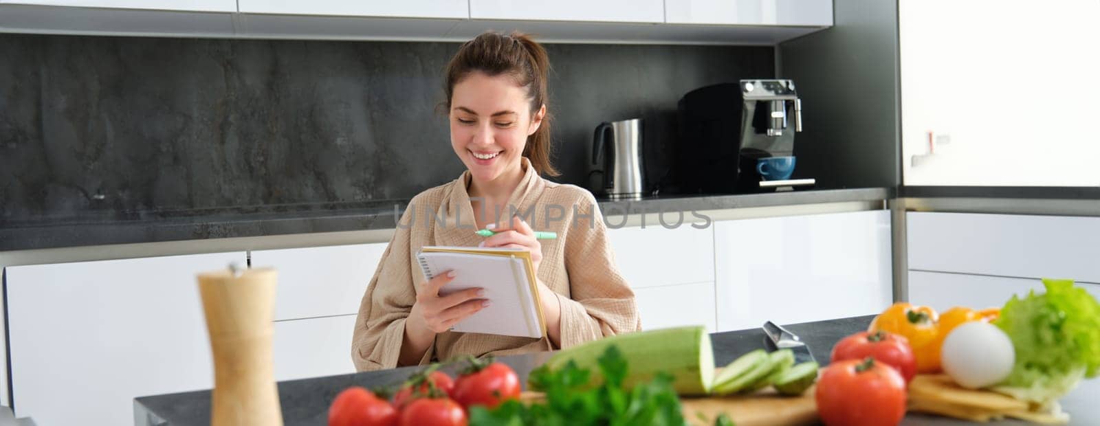 Portrait of beautiful, smiling young woman making list of meals, writing down recipe, sitting in the kitchen with vegetables, doing house errands.