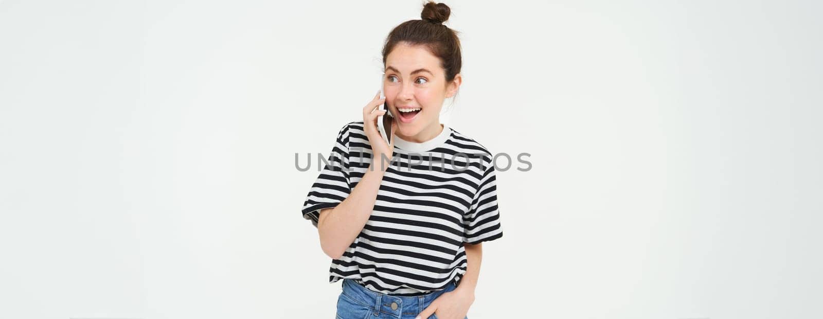 Portrait of enthusiastic young woman talking on mobile phone, chatting with friend, reacts to surprising news received by telephone, white background.