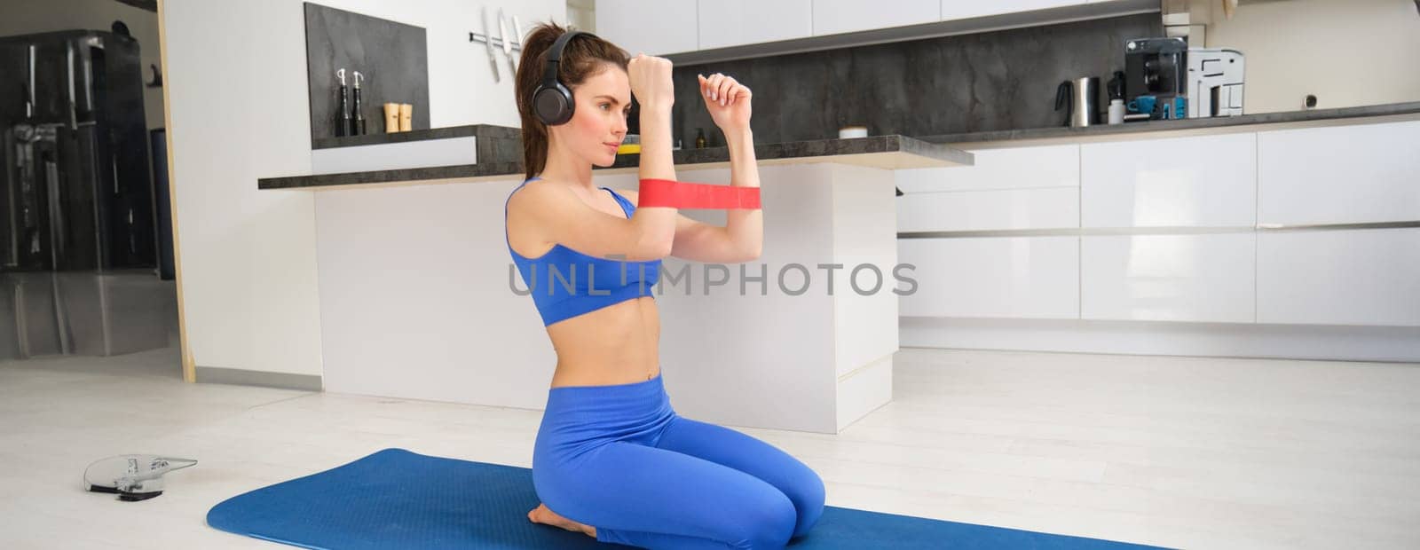 Young woman doing fitness workout at home, using elastic resistance band on arms, muscle exercises, sitting on rubber mat in living room.