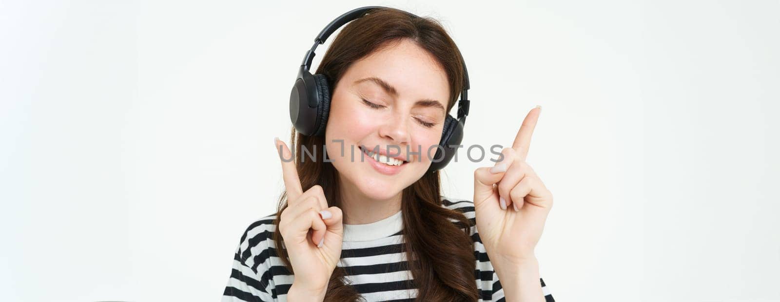 Portrait of happy student girl, listening music in headphones, dancing and pointing fingers at copy space, promo text, isolated against white background.