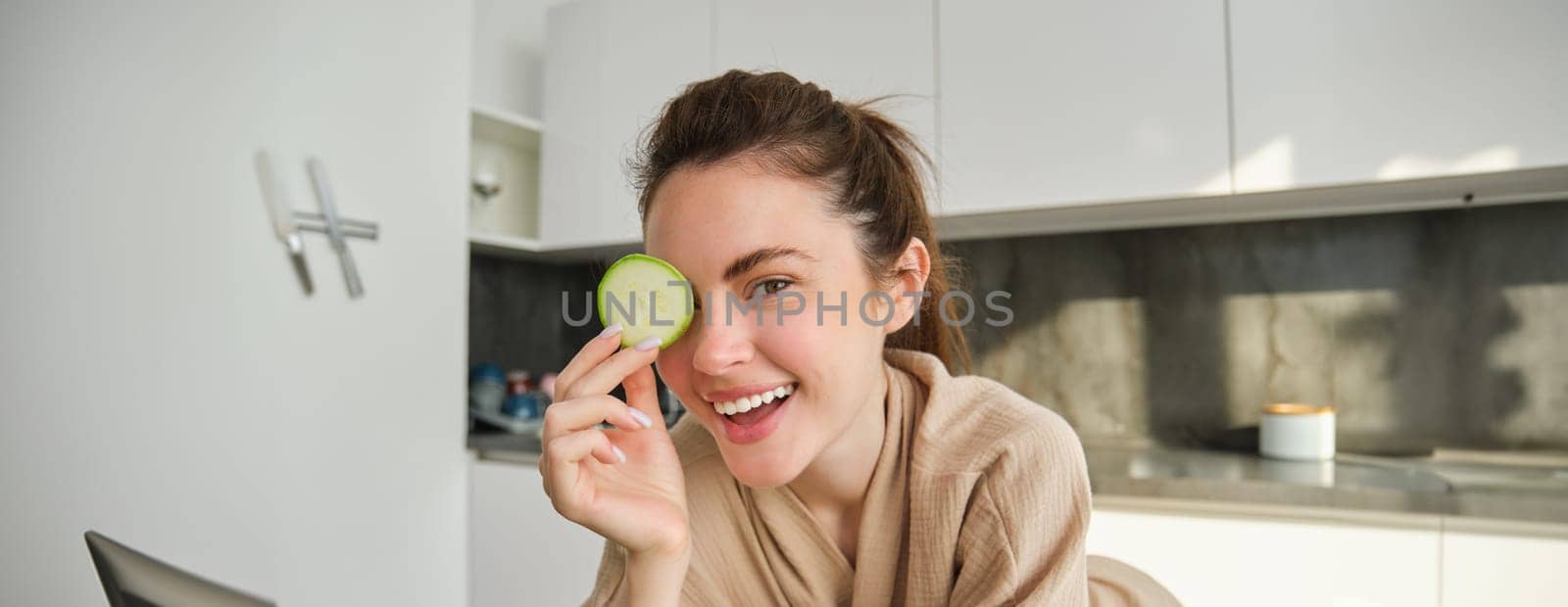 Portrait of happy, smiling young woman in the kitchen, cooking, chopping zucchini, holding vegetables and looking happy, preparing vegan food meal at home by Benzoix