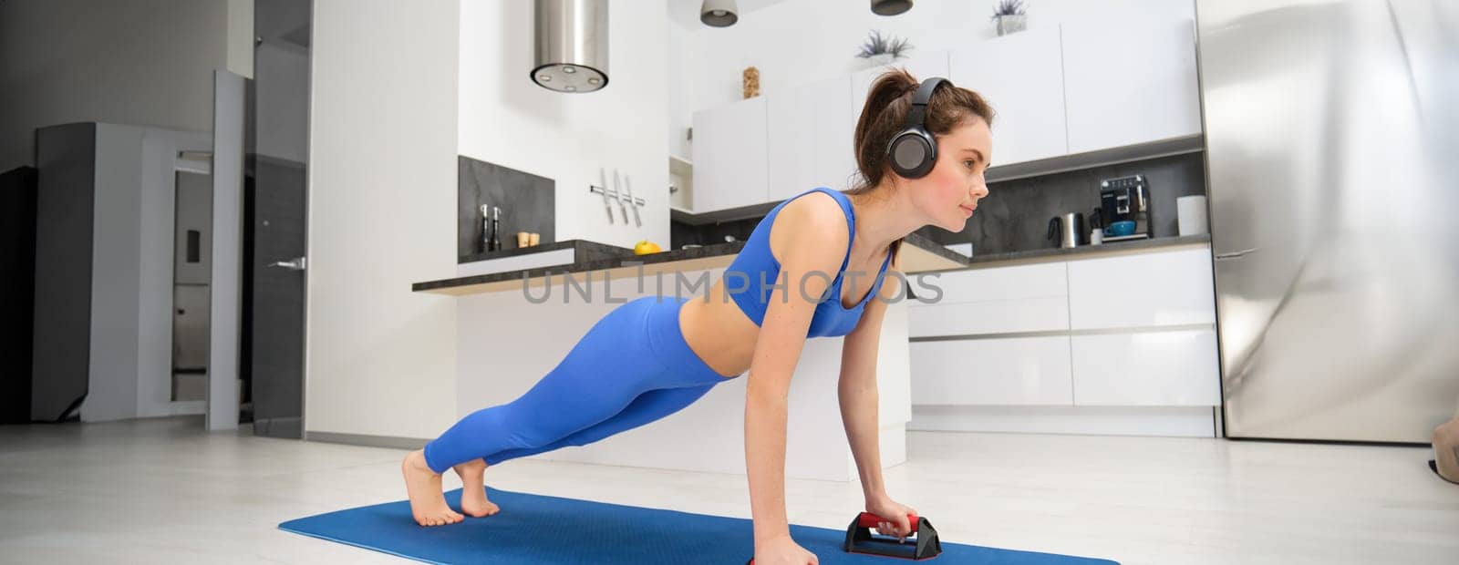 Portrait of woman in wireless headphones, doing workout from home, standing in plank with sports equipment, listening music, looking confident.