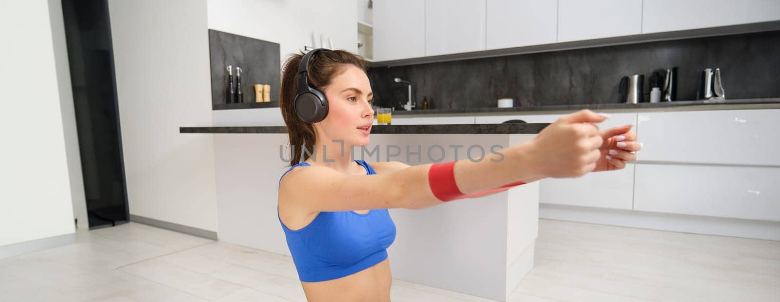 Image of hardworking sportswoman, girl doing fitness exercises at home, listening music in headphones, stretching elastic resistance band with arms, workout in living room.