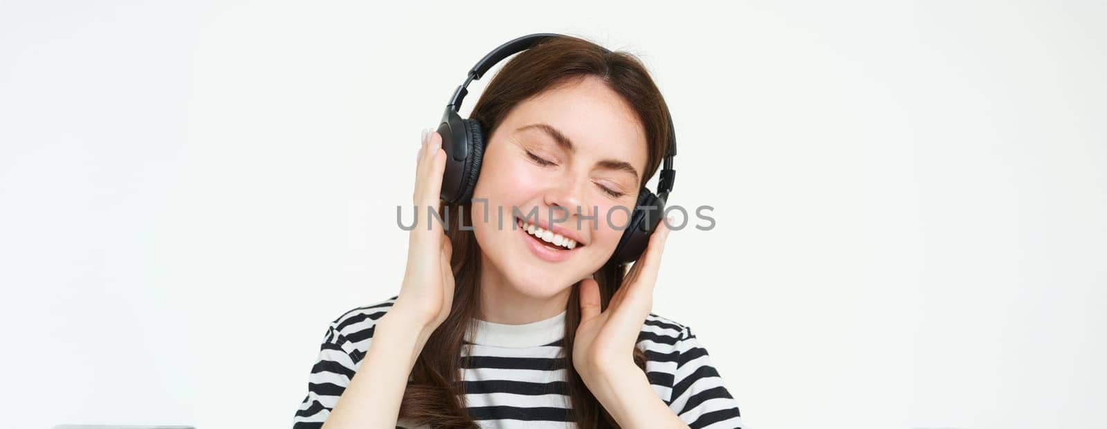 Portrait of beautiful woman in wireless headphones, listening music, using earphones, smiling at camera, standing over white background.