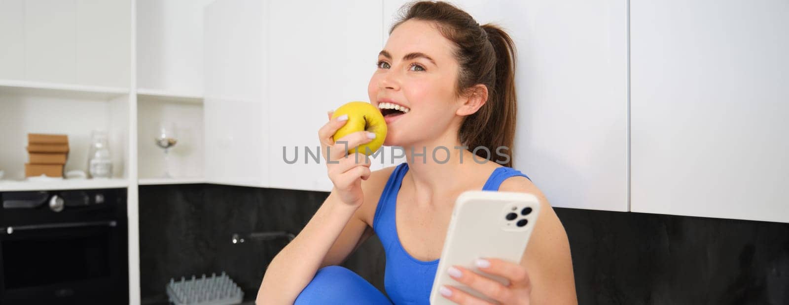 Portrait of stylish, young fitness woman, eating an apple and using mobile phone, holding smartphone, wearing sportswear.