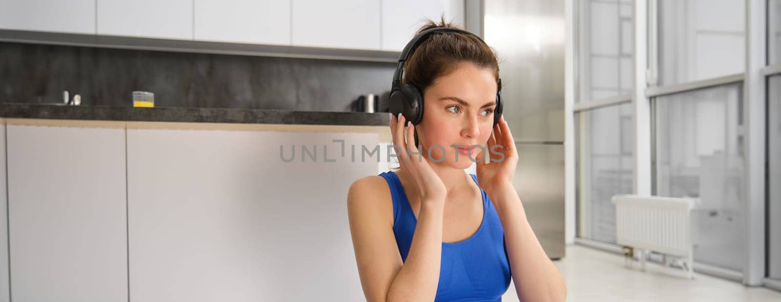 Image of fitness girl doing sports at home, puts on wireless headphones for music during workout training session, wearing blue sportsbra.