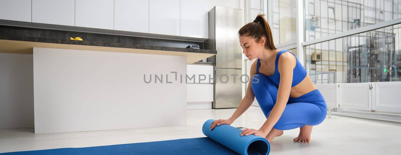 Image of young woman preparing living room for meditation, yoga exercises, unwrapping a mat on living room floor, wearing sportswear.