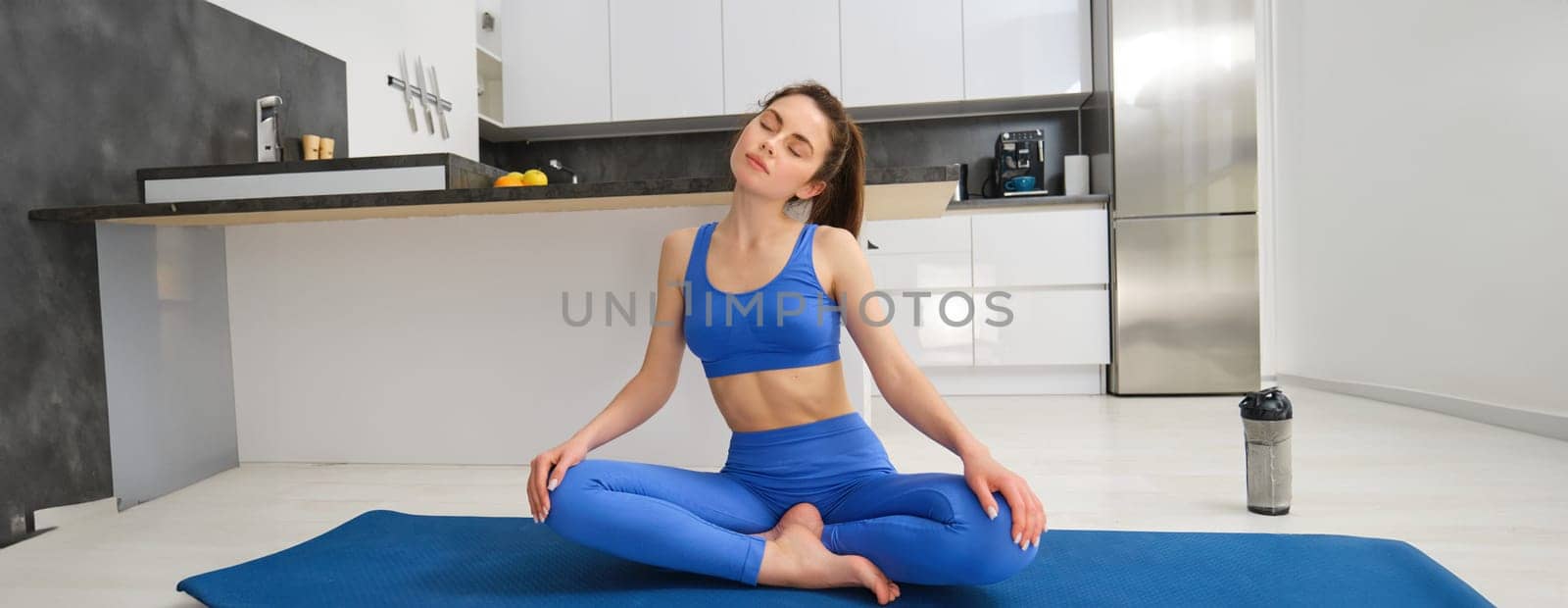 Portrait of young sportswoman, stretching her neck, warm-up before yoga exercises, doing fitness workout on rubber mat, wearing blue sportsbra and leggings.