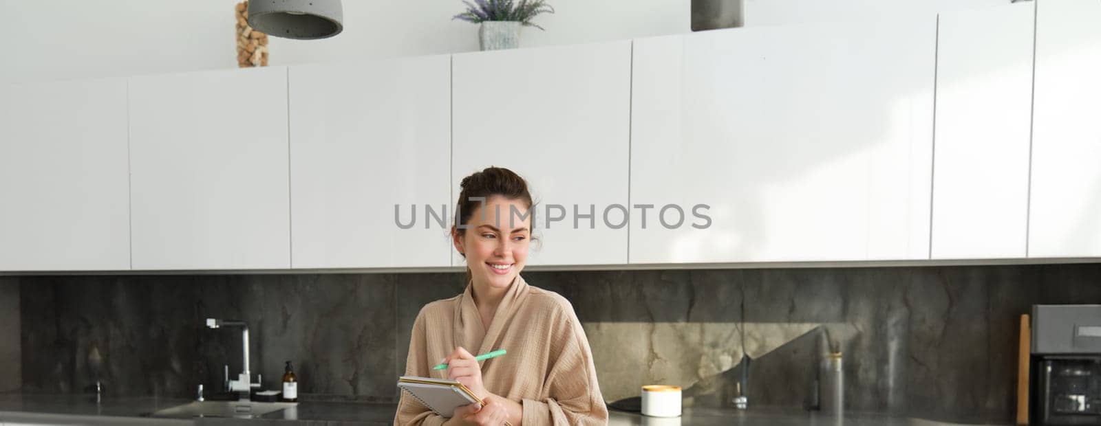 Vertical shot of brunette woman in bathrobe, cooking in the kitchen, writing down recipe in notebook, standing near chopping board with vegetables, making salad and smiling by Benzoix