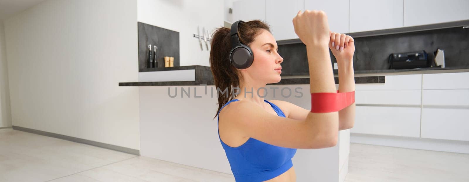 Focused sportswoman workout at home, using elastic resistance band on arms, stretching exercises in living room, aerobics training.