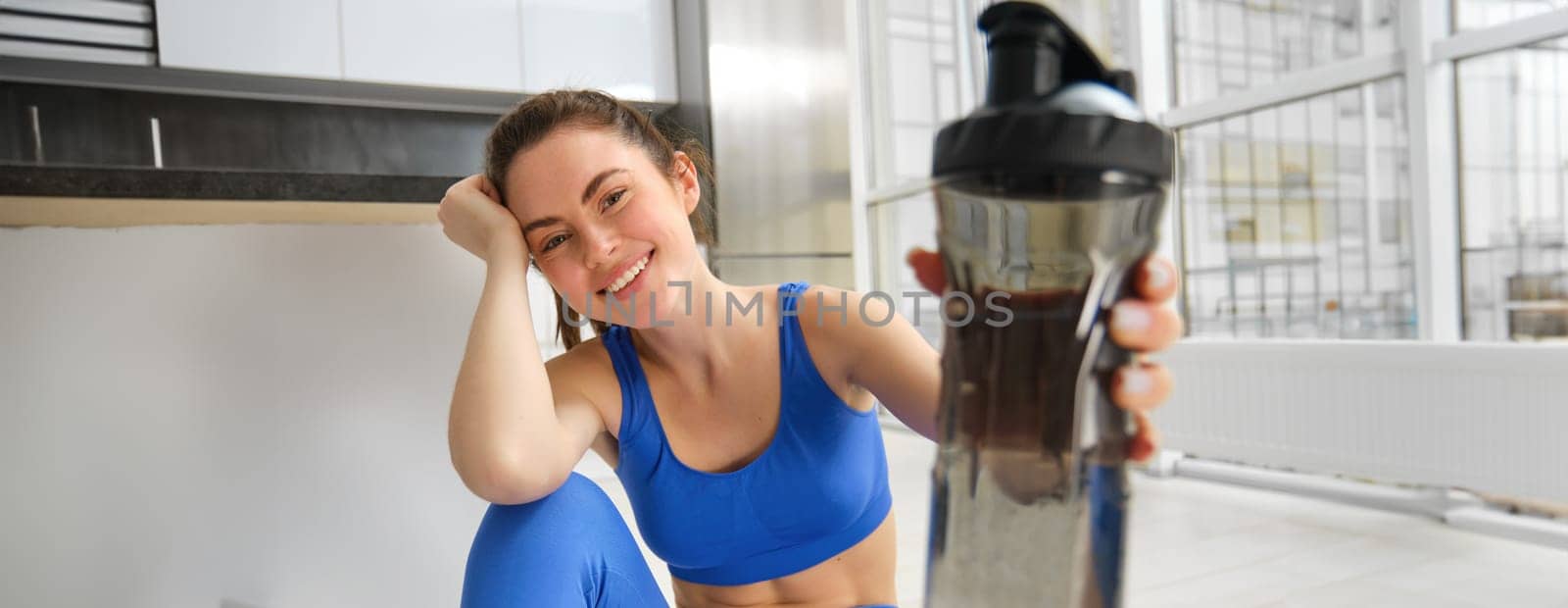 Young woman working out from home, drinking water during her sport training session in living room, sitting on yoga mat by Benzoix