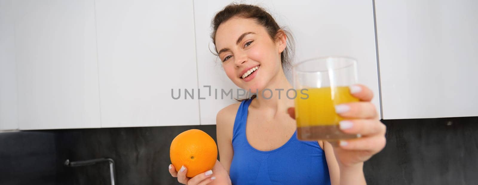 Portrait of smiling fitness woman, offering orange juice, holding fruit and a glass in hands, posing in kitchen in activewear.