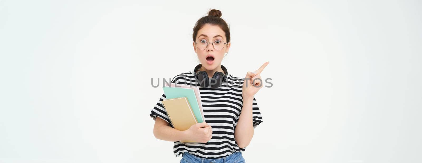 Portrait of girl gasps in surprised, points left with amazed face. Shocked student shows something, pointing finger at banner, holding notebooks, white background.