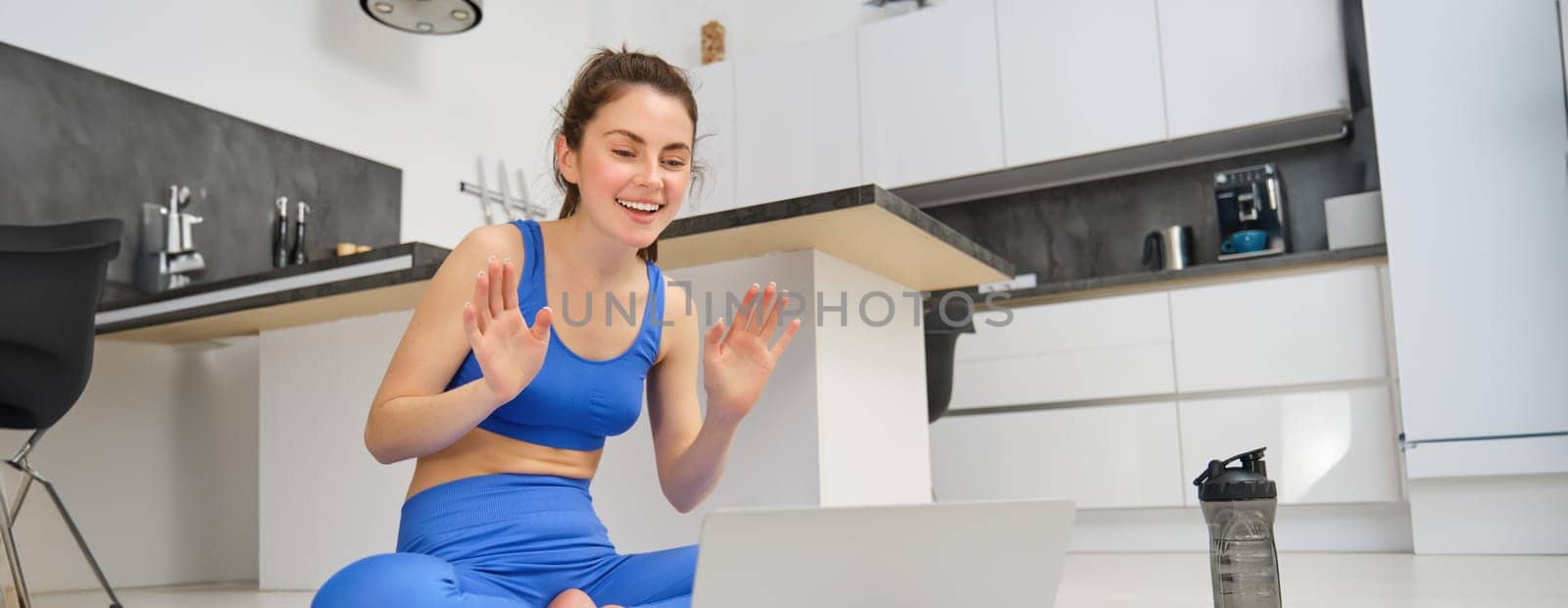Portrait of fit and healthy, female fitness instructor sitting on floor at home, talking to laptop, teaching online class yoga or workout training.