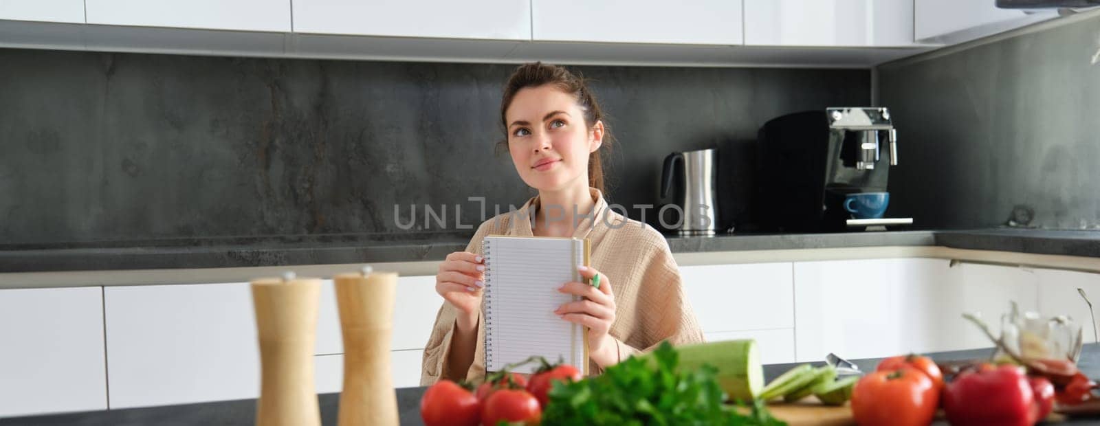 Portrait of happy young woman writes down menu for dinner, sits in the kitchen near vegetables, makes grocery list for shopping, poses in the bathrobe by Benzoix