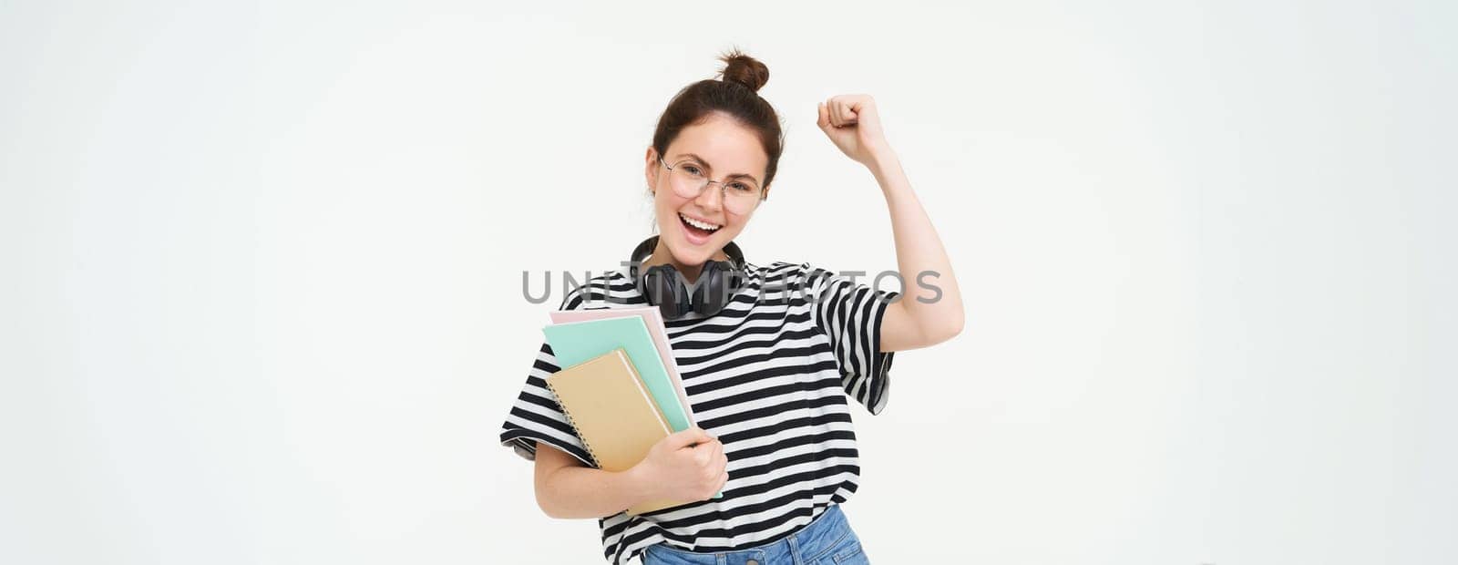 Portrait of young woman in glasses, wearing headphones over neck, holding planner, notebooks and study material, cheering, rooting for you, celebrating, standing over white background.