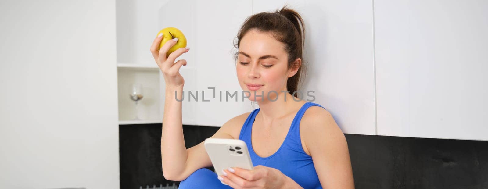 Portrait of brunette fitness woman, eating an apple, holding smartphone, using mobile phone app while having healthy fruit snack in kitchen.