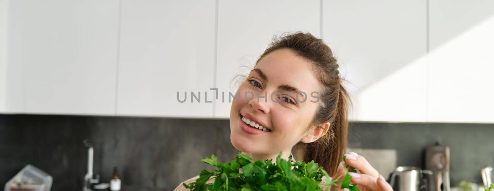 Portrait of beautiful woman cooking, holding fresh parsley, adding herbs while making salad, healthy meal in the kitchen by Benzoix