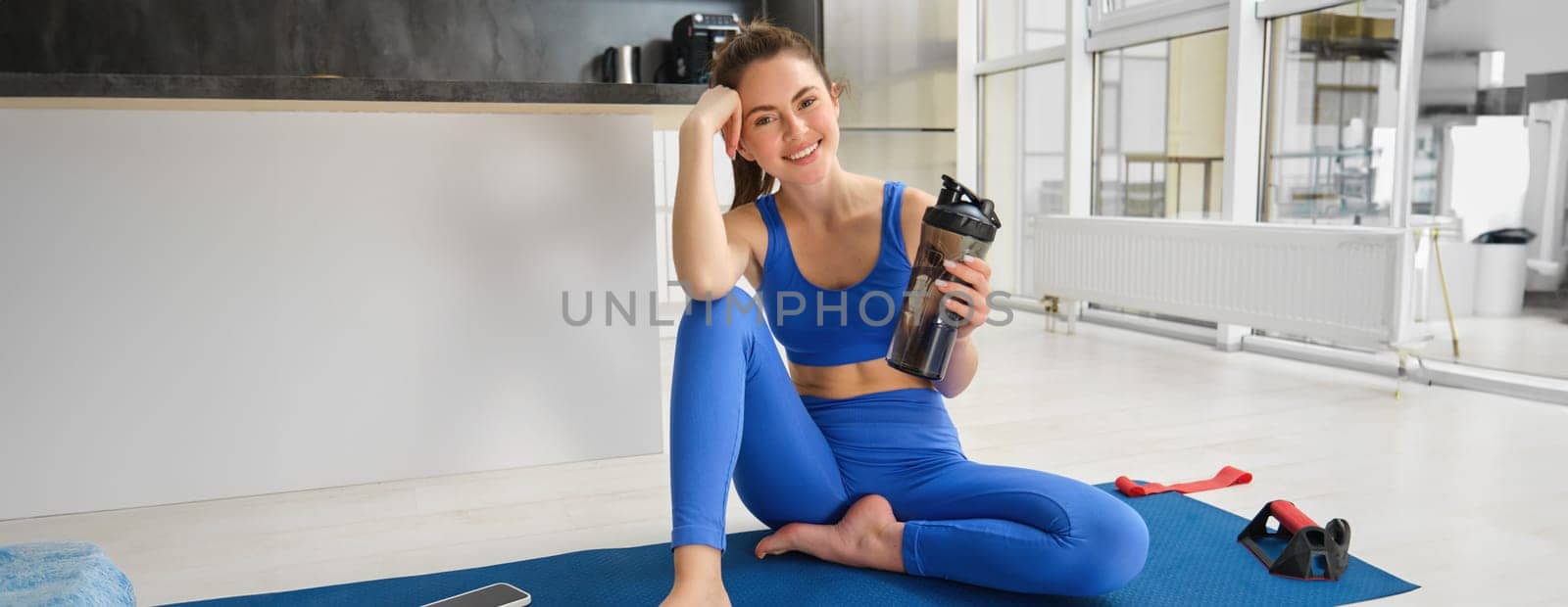 Portrait of smiling fitness instructor, drinks water from bottle, sits on yoga mat, workout from home.