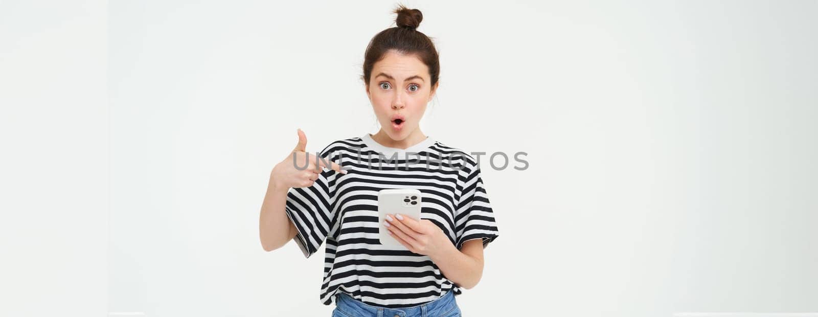 Portrait of impressed woman, drops jaw, points at her smartphone, sees amazing news online on mobile phone, isolated over white background by Benzoix