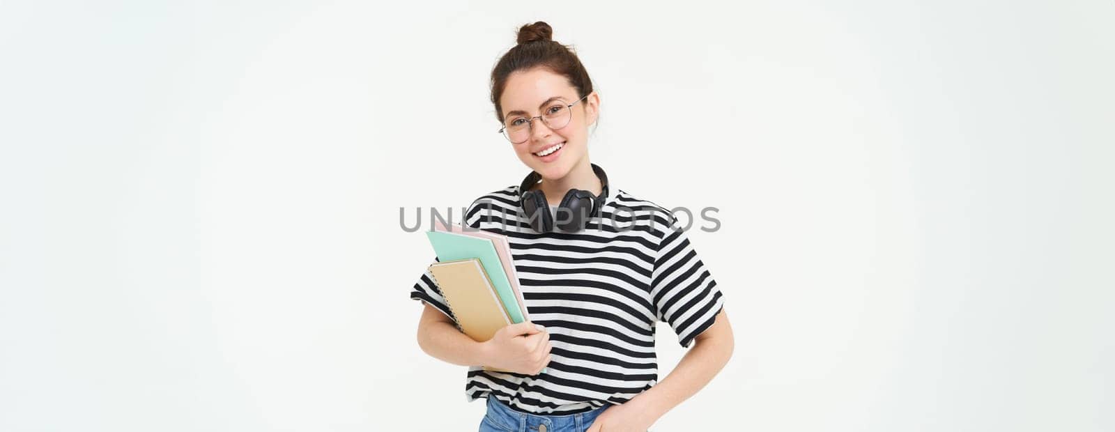 Young woman with notebooks, books and study material, posing over white background, wears headphones over neck.