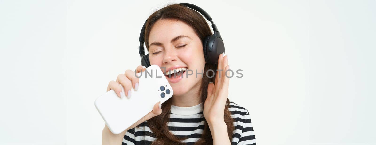 Portrait of happy girl playing karaoke app, singing into smartphone microphone, wearing wireless headphones, white background by Benzoix