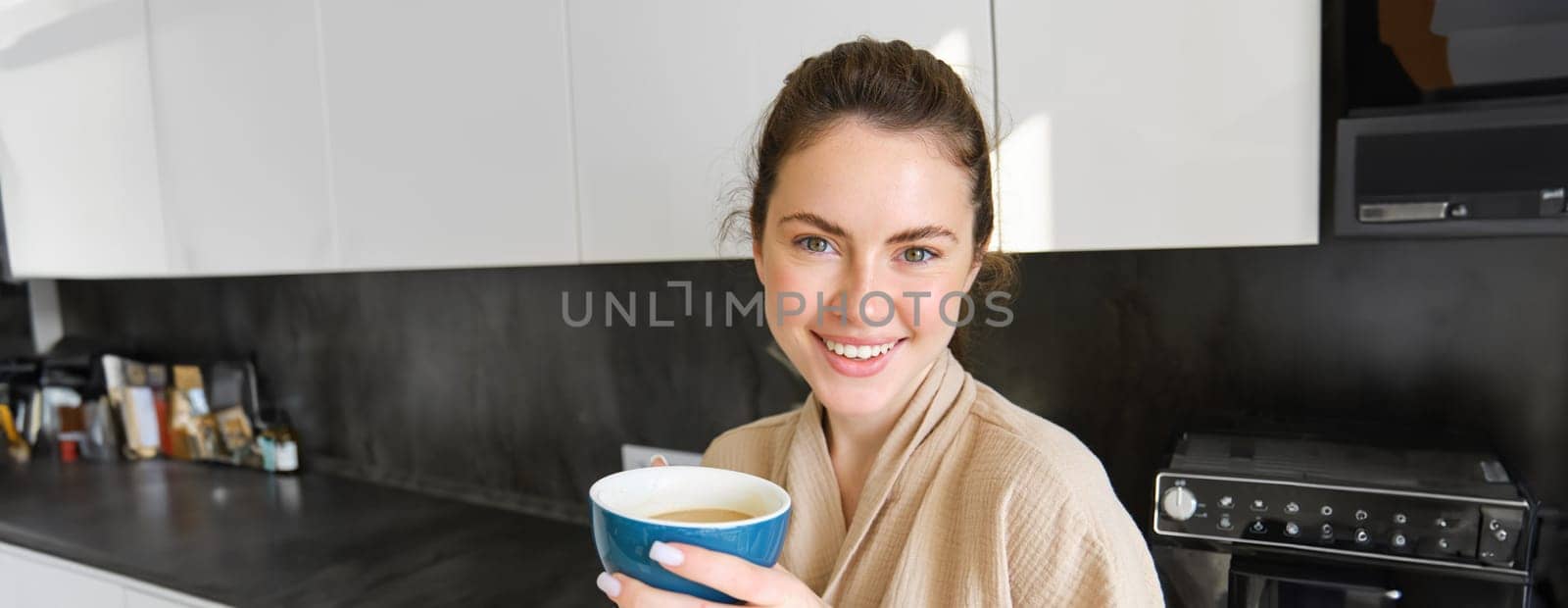 Good-looking young woman with cup of coffee, posing in the kitchen, smiling at camera, enjoys her morning at home, wearing cosy bathrobe.