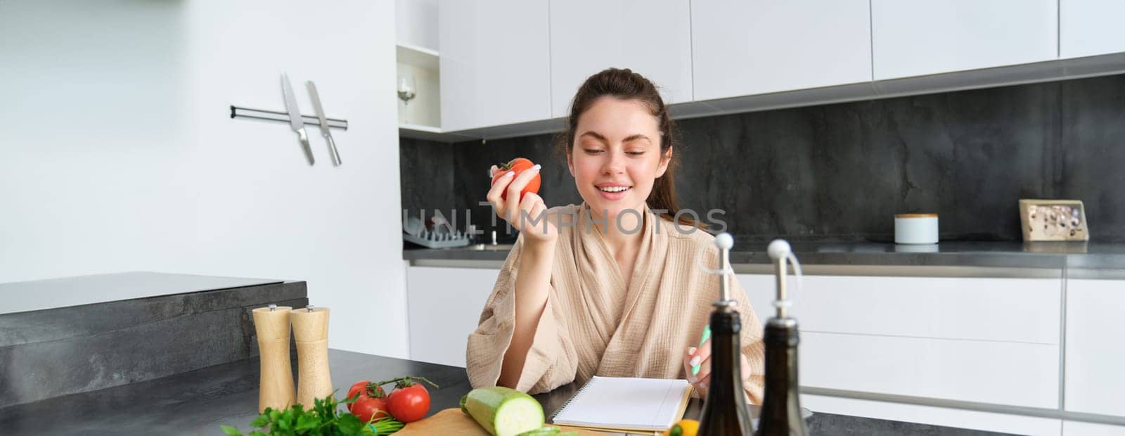Portrait of beautiful smiling woman, writing her healthy menu, eating tomato while cooking, making grocery list, sitting in the kitchen by Benzoix
