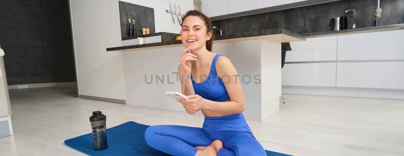 Portrait of sportswoman using sport app while doing workout, yoga exercises at home, sits on mat with water bottle, smiling and looking happy by Benzoix