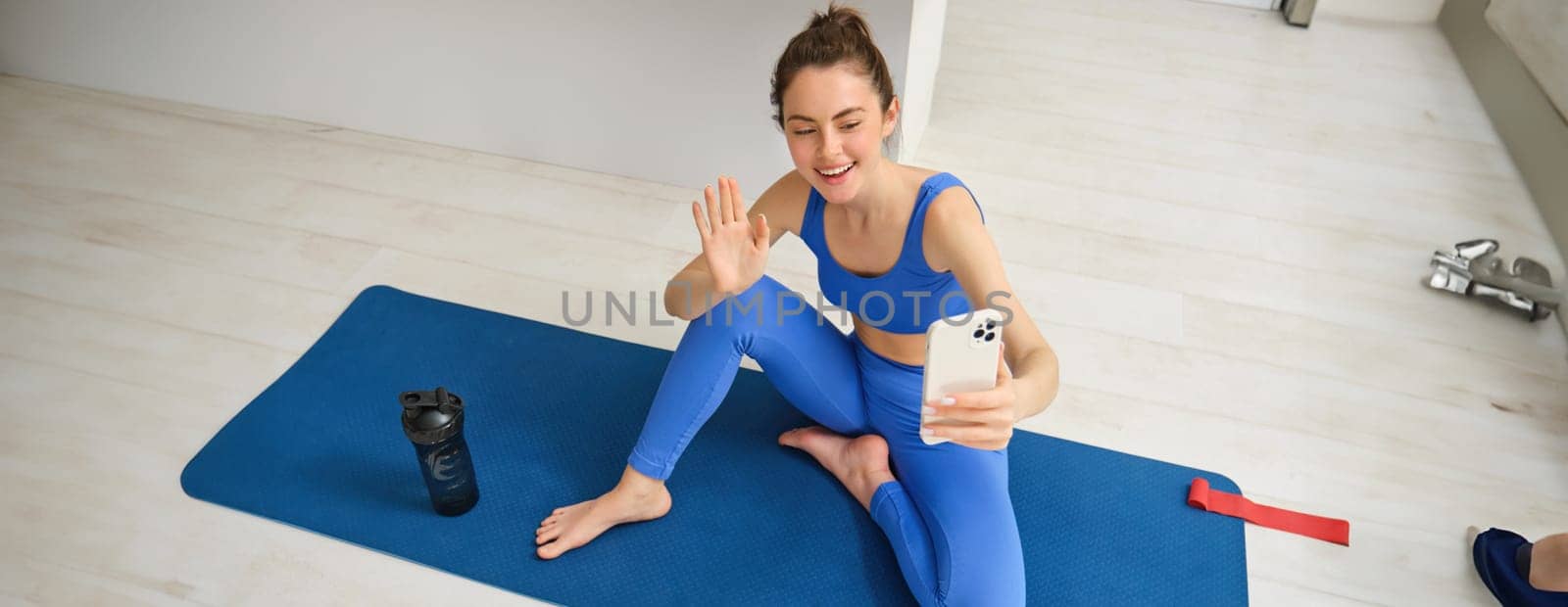 Portrait of young styling fitness girl doing workout from home, taking selfie and video for social media, gym instructor records her training session indoors.