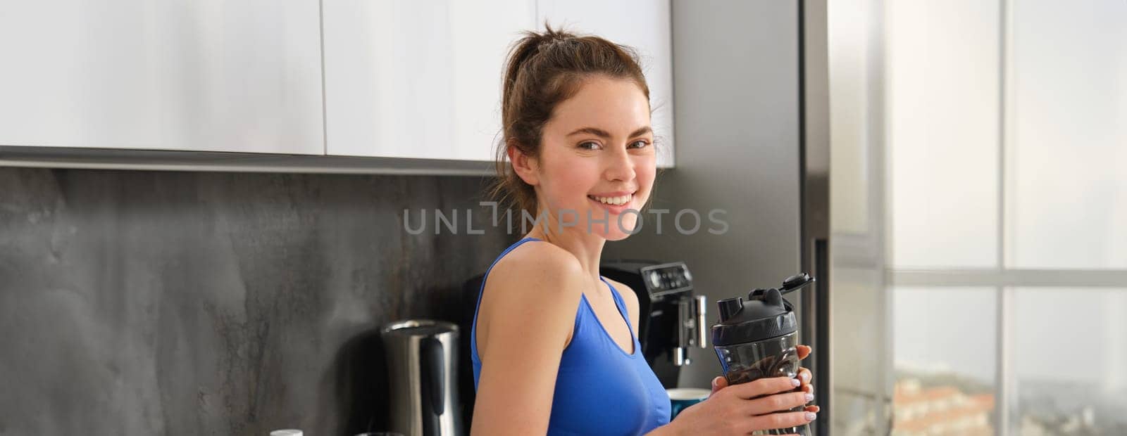 Image of young fitness instructor, woman in sportsbra and leggings, holding water bottle, drinking after workout, standing in kitchen.