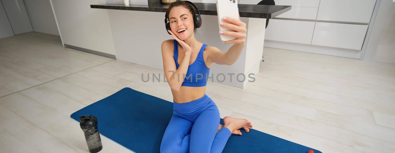 Beautiful and stylish sportswoman, posing for selfie, listens music in wireless headphones, holds smartphone, sits on fitness mat, workout from home.