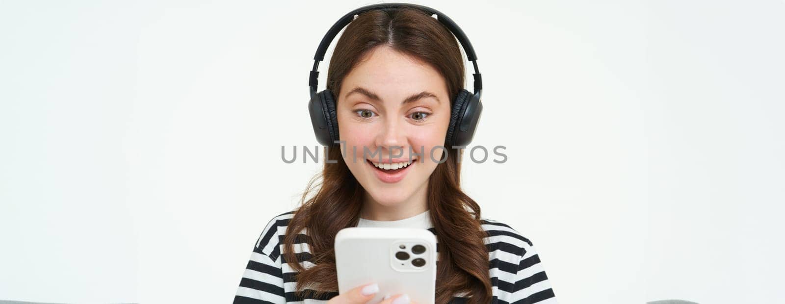 Young woman with surprised face, looking at smartphone screen, reacts amazed, looks with happiness at mobile phone, standing in wireless headphones, white background.