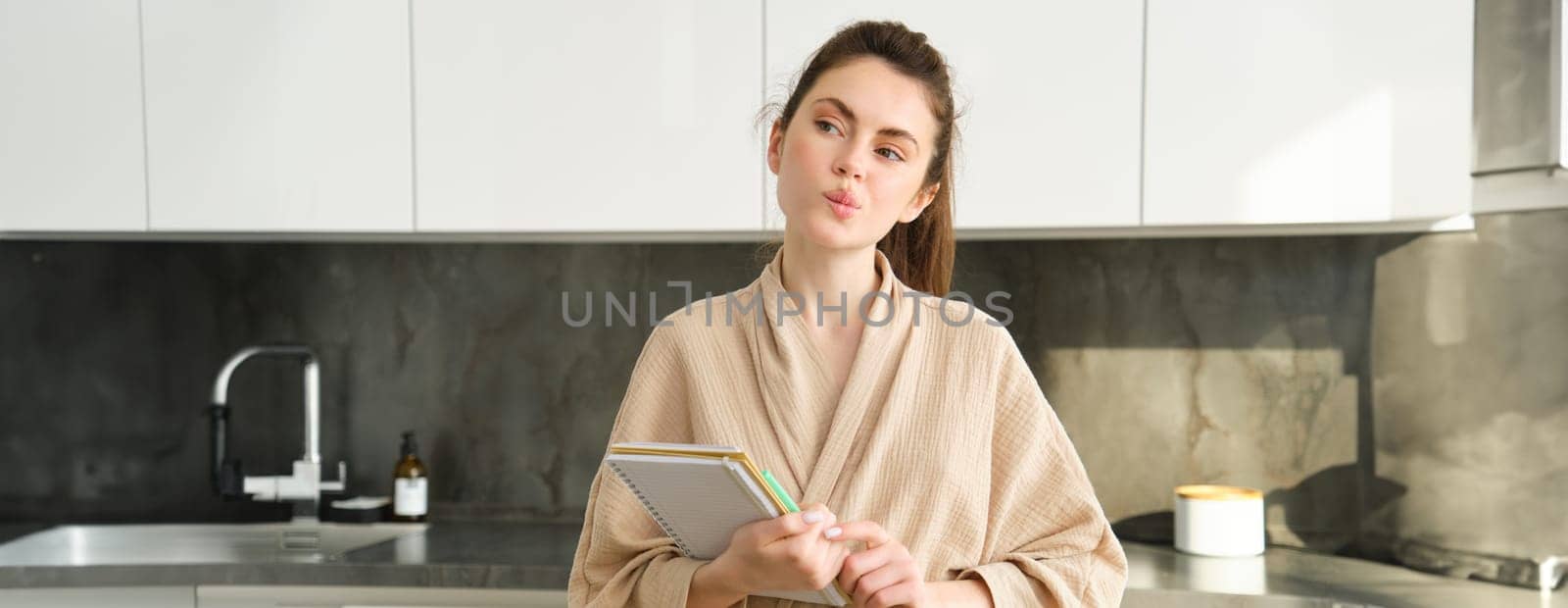 Portrait of beautiful modern woman, writing down grocery list, meal ideas in notebook, standing in the kitchen, wearing cosy bathrobe by Benzoix