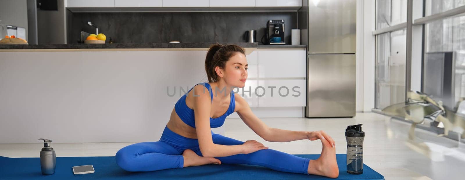 Image of young fit woman, does gymanstics at home, stretches her legs on floor in living room, does fitness workout, yoga warm-up exercises by Benzoix