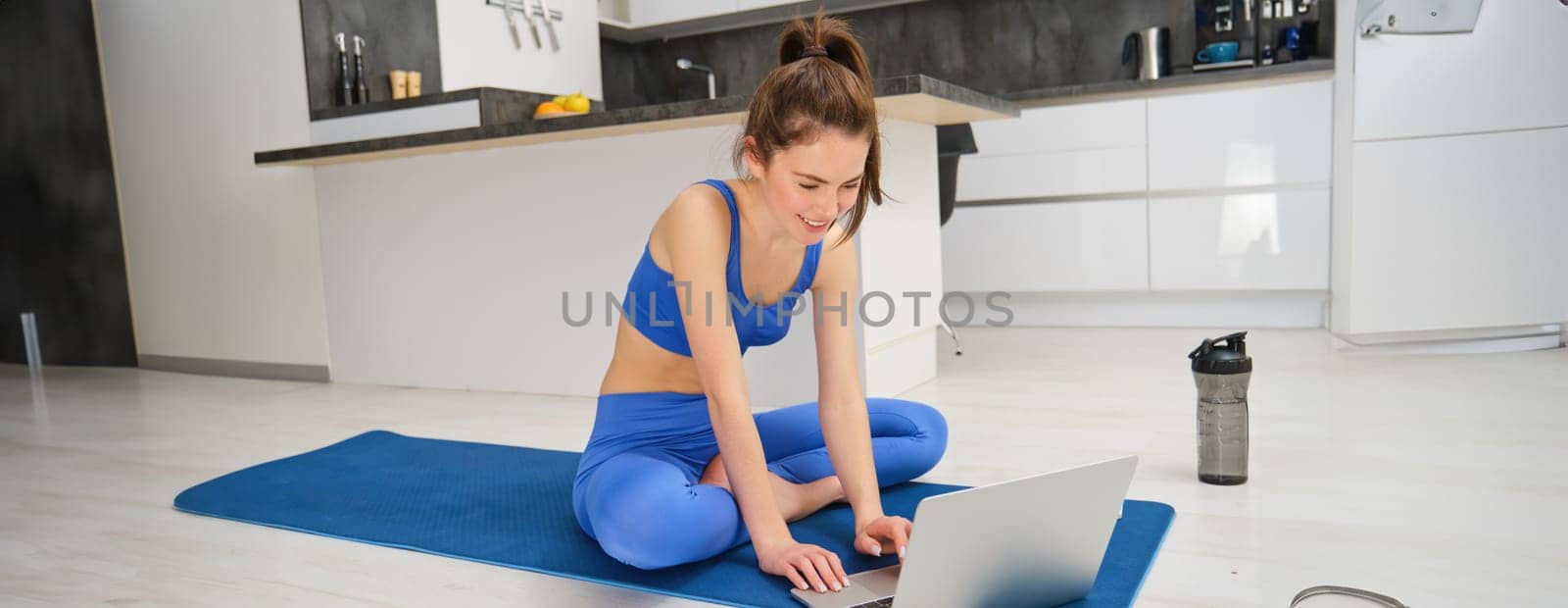 Image of young fitness instructor using laptop for online workout training, showing fitness exercises, talking to client, sitting on yoga rubber mat in sportswear.