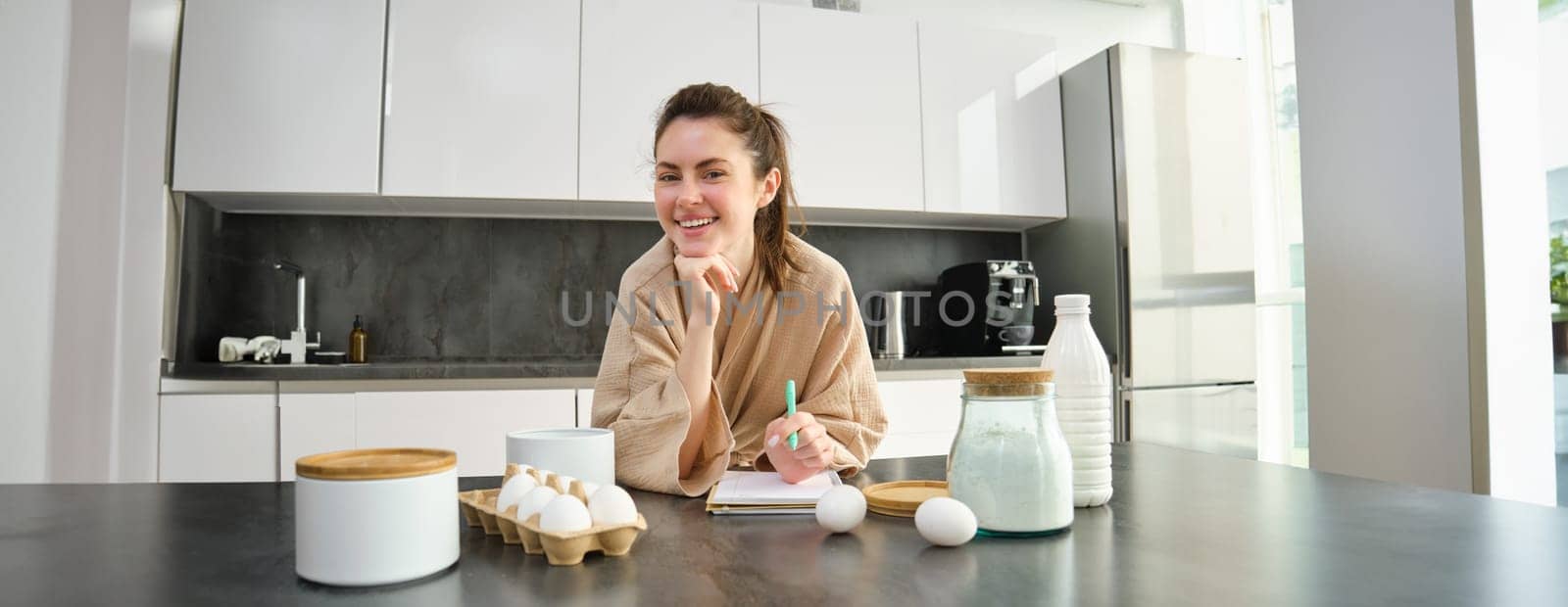 Attractive young cheerful girl baking at the kitchen, making dough, holding recipe book, having ideas.