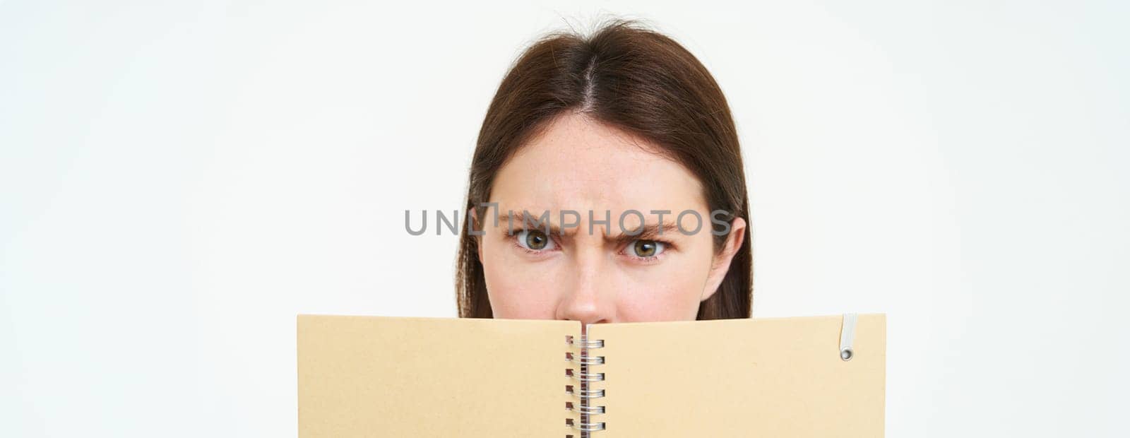 Woman with confused face looking at camera, frowning, holding notebook, reading planner or personal diary, standing over white background.