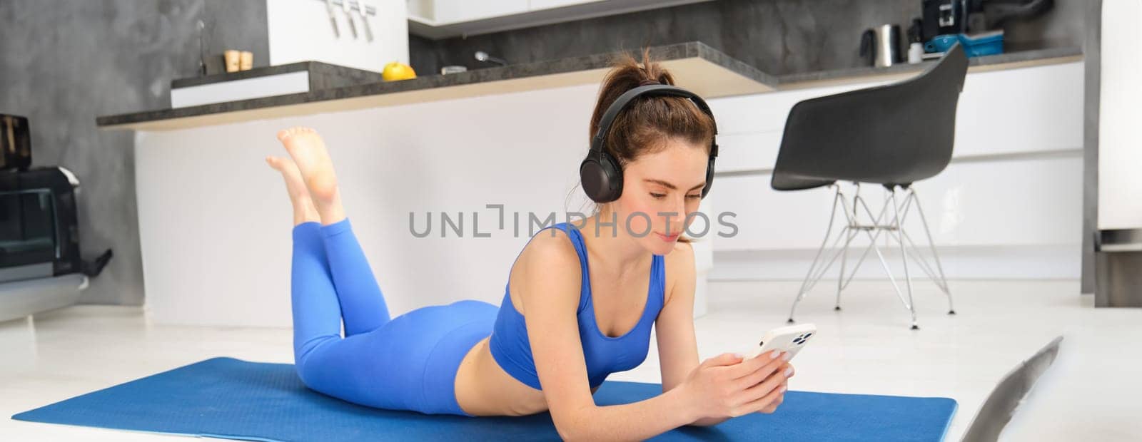 Image of young woman watches sport gym tutorials on smartphone, laying on yoga mat, doing fitness workout at home, wearing blue leggings and sportsbra.