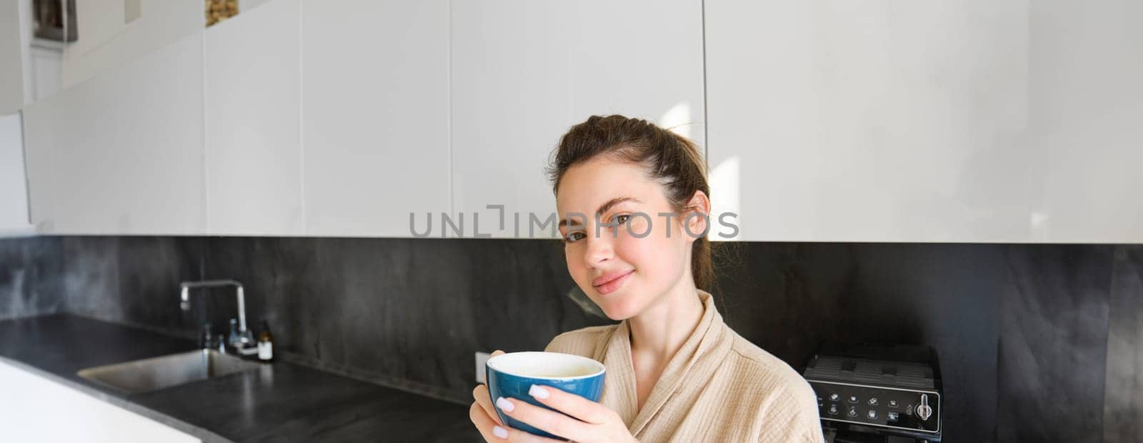 Portrait of good-looking young woman, drinking coffee in the kitchen, enjoying her morning routine and smiling at the camera.